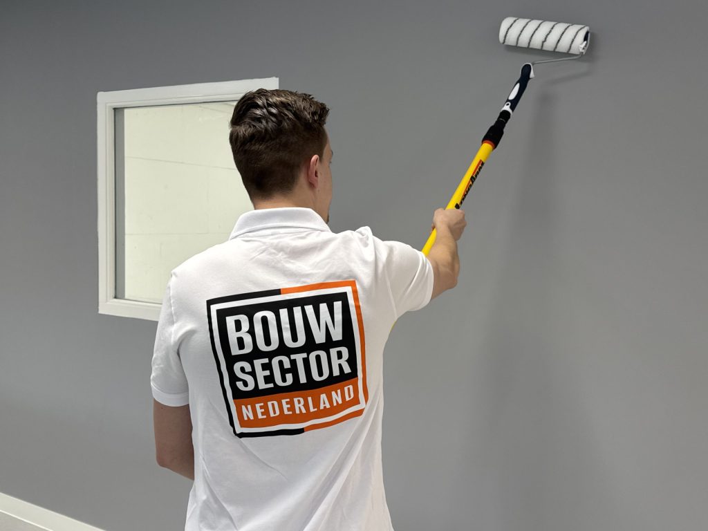 Schilder in witte werkpolo rolt een grijze muur vlak naast een wit gelakt raamkozijn